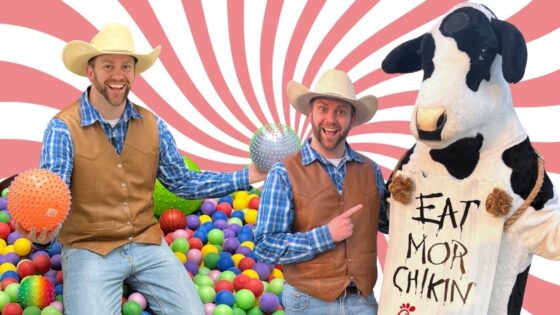 Two men in cowboy vests and hats pose in a colorful ball pit with a life-size cow mascot holding a sign that says 'EAT MOR CHIKIN' against a pink swirl background.