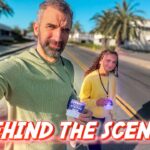 Two people walk on a sunny street wearing lanyards with visitor badges; bold 'Behind the Scenes' text overlays the bottom of the thumbnail.