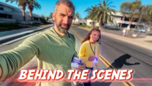 Two people walk on a sunny street wearing lanyards with visitor badges; bold 'Behind the Scenes' text overlays the bottom of the thumbnail.