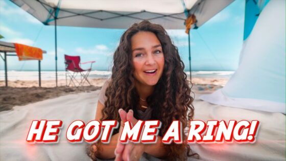 Woman on a sunny beach under a canopy, smiling, with a bold red text overlay reading 'HE GOT ME A RING!'