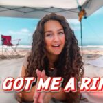Woman with long curly hair on a sunny beach under a canopy, smiling with hands together, bold caption: 'HE GOT ME A RING!'