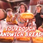 Woman presenting a large sourdough loaf to a group of kids gathered around a kitchen island.