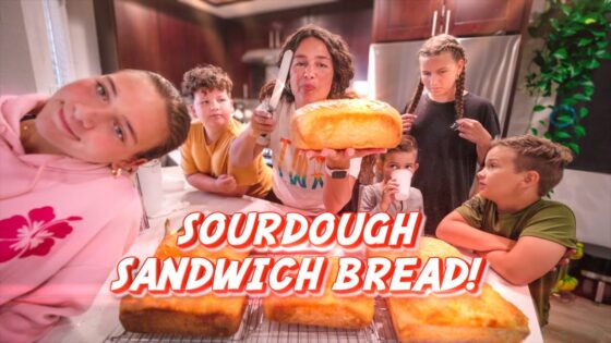 Woman presenting a large sourdough loaf to a group of kids gathered around a kitchen island.