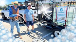 Two workers in hats pose beside a trailer-mounted pressure washer advertising exterior cleaning services in a parking lot with a white pickup nearby.