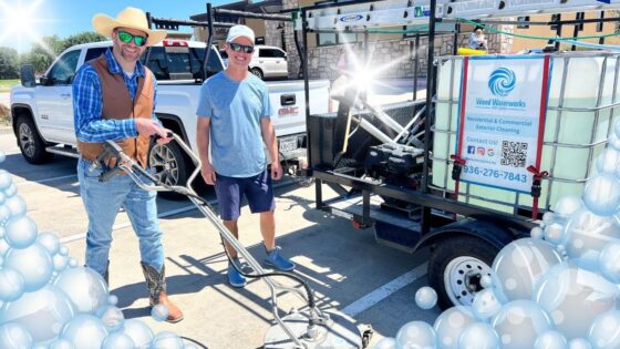 Two workers in hats pose beside a trailer-mounted pressure washer advertising exterior cleaning services in a parking lot with a white pickup nearby.