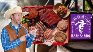 Man in a cowboy hat and blue plaid shirt points excitedly at a red tray of smoked meats (ribs, brisket, turkey) in a BBQ setup with a purple Bar-A-BBQ sign from Montgomery, Texas.