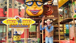 Man in a cowboy hat and vest points at a colorful Billy Beez play area sign inside an indoor kids' attraction park.