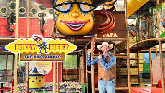 Man in a cowboy hat and vest points at a colorful Billy Beez play area sign inside an indoor kids' attraction park.
