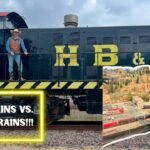 A cowboy in a brown vest and hat stands on a dark locomotive with yellow lettering; a neon-framed sign reads 'REAL TRAINS VS. MODEL TRAINS!!!' and a model railroad layout is shown on the right with another man pointing.