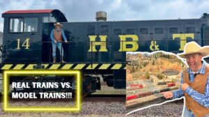 A cowboy in a brown vest and hat stands on a dark locomotive with yellow lettering; a neon-framed sign reads 'REAL TRAINS VS. MODEL TRAINS!!!' and a model railroad layout is shown on the right with another man pointing.