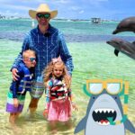 Man with two children in life jackets posing in shallow, clear water as dolphins jump nearby on a sunny beach day with boats on the horizon.