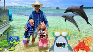 Man with two children in life jackets posing in shallow, clear water as dolphins jump nearby on a sunny beach day with boats on the horizon.