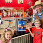 Smiling man in a cowboy hat and brown vest poses with two kids at an ice cream shop counter.