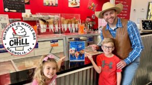 Smiling man in a cowboy hat and brown vest poses with two kids at an ice cream shop counter.