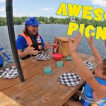 Group of kids in life jackets cheer around a picnic table on a lake, with checkered plates and a wicker basket nearby.