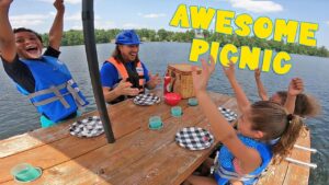 Group of kids in life jackets cheer around a picnic table on a lake, with checkered plates and a wicker basket nearby.