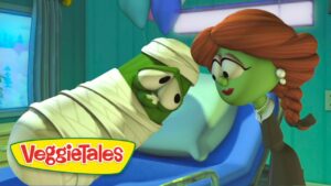 VeggieTales scene: a green mummy-like character wrapped in bandages on a hospital bed, leaning toward a woman with red braided hair.