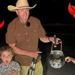 Man wearing a tan cowboy hat and brown shirt at night on a beach, holding a lantern with two children beside him, red cartoon crabs on the sides.