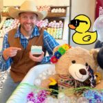 Man in a cowboy hat and brown vest smiles while pointing to a bar of soap in a colorful soap shop display, with a teddy bear in a tub adorned with flowers and balloons nearby.