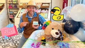 Man in a cowboy hat and brown vest smiles while pointing to a bar of soap in a colorful soap shop display, with a teddy bear in a tub adorned with flowers and balloons nearby.