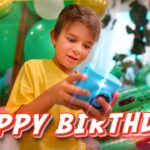Young boy in a yellow shirt celebrates a birthday, holding a blue toy with green balloons and party decorations behind him; a 'HAPPY BIRTHDAY' banner is across the bottom.