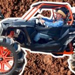 Off-road buggy with bright red wheels on rocky dirt, driver wearing a cowboy hat and blue plaid shirt in the cockpit.