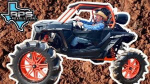 Off-road buggy with bright red wheels on rocky dirt, driver wearing a cowboy hat and blue plaid shirt in the cockpit.