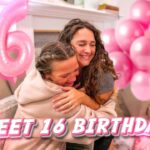 Two people hug in a pink‑balloon decorated living room for a Sweet 16 birthday celebration.