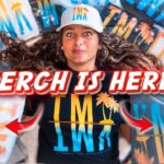 Person lying among colorful wrapped merch bags with a bold 'MERCH IS HERE!' overlay text, promotional thumbnail avoid reference to identity.