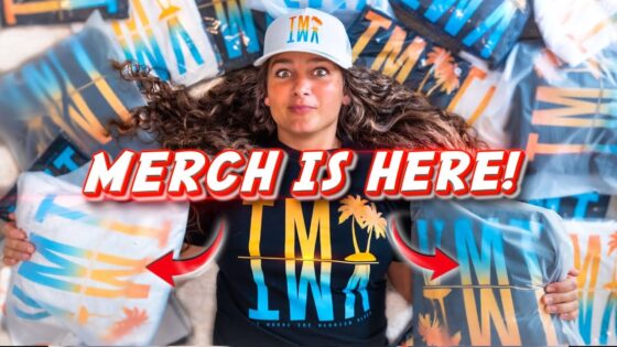 Person lying among colorful wrapped merch bags with a bold 'MERCH IS HERE!' overlay text, promotional thumbnail avoid reference to identity.