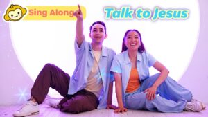 Two smiling hosts sit on the floor in pastel outfits, inviting viewers to sing along and talk to Jesus.
