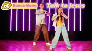 Two young dancers perform a joyful dance on a stage with purple neon vertical lights and 'Dance Along'/'Talk to Jesus' banners.