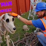 Man in an orange safety vest and blue helmet pets a life-size skeleton horse in a Halloween display by a red wall.