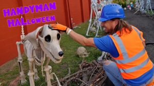 Man in an orange safety vest and blue helmet pets a life-size skeleton horse in a Halloween display by a red wall.