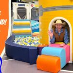 Man in a cowboy hat waves from the entrance of a bright yellow play ship; colorful cushions, a ball pit, and an orange banner reading 'Kiddie Cosmos Playground' with a cartoon cowboy.
