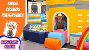 Man in a cowboy hat waves from the entrance of a bright yellow play ship; colorful cushions, a ball pit, and an orange banner reading 'Kiddie Cosmos Playground' with a cartoon cowboy.