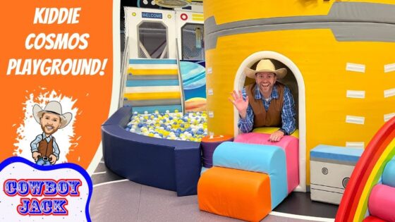 Man in a cowboy hat waves from the entrance of a bright yellow play ship; colorful cushions, a ball pit, and an orange banner reading 'Kiddie Cosmos Playground' with a cartoon cowboy.