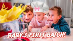 Three kids in a kitchen watch a mixer as yellow batter splashes, with a baby wearing a chef hat and pink bow; the image is overlaid with the text "BABY'S FIRST BATCH!"