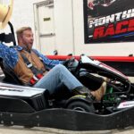 Man in a go-kart raising a straw hat with an excited expression indoors, wearing a blue plaid shirt and brown vest.
