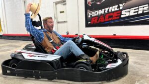 Man in a go-kart raising a straw hat with an excited expression indoors, wearing a blue plaid shirt and brown vest.