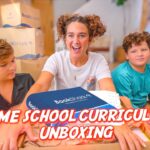 Three kids unbox a BookShark home-school curriculum box in a living room, with 'HOME SCHOOL CURRICULUM UNBOXING' text visible in the foreground.