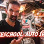 Man in a garage holding a bottle of Valvoline motor oil next to a car, with a bold red 'HOMESCHOOL AUTO SHOP?' overlay in the foreground.