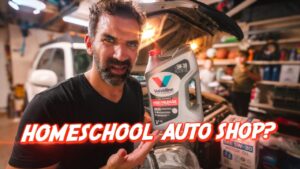 Man in a garage holding a bottle of Valvoline motor oil next to a car, with a bold red 'HOMESCHOOL AUTO SHOP?' overlay in the foreground.