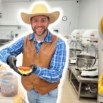 Smiling man in a cowboy hat and brown vest builds a sandwich in a commercial bakery kitchen, with mixers and cookware in the background.