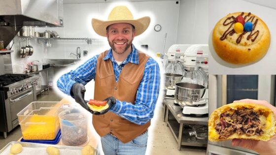 Smiling man in a cowboy hat and brown vest builds a sandwich in a commercial bakery kitchen, with mixers and cookware in the background.