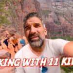 Man with beard taking a selfie on a rocky canyon trail, with several kids hiking behind him under red cliffs.