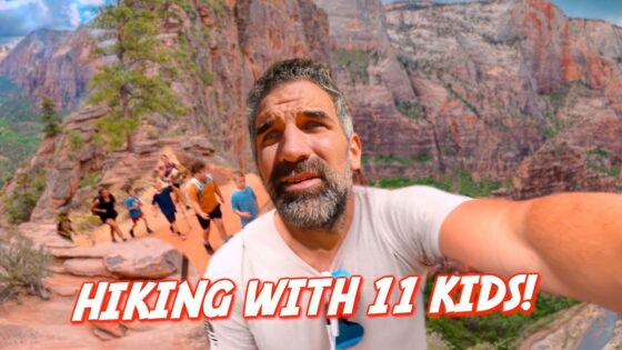 Man with beard taking a selfie on a rocky canyon trail, with several kids hiking behind him under red cliffs.