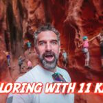 Selfie of a bearded man taking a photo inside a red canyon while several children wear helmets and climb the rock walls, with the bold caption 'Exploring with 11 kids!' across the bottom.