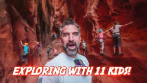 Selfie of a bearded man taking a photo inside a red canyon while several children wear helmets and climb the rock walls, with the bold caption 'Exploring with 11 kids!' across the bottom.