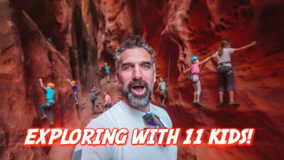 Selfie of a bearded man taking a photo inside a red canyon while several children wear helmets and climb the rock walls, with the bold caption 'Exploring with 11 kids!' across the bottom.
